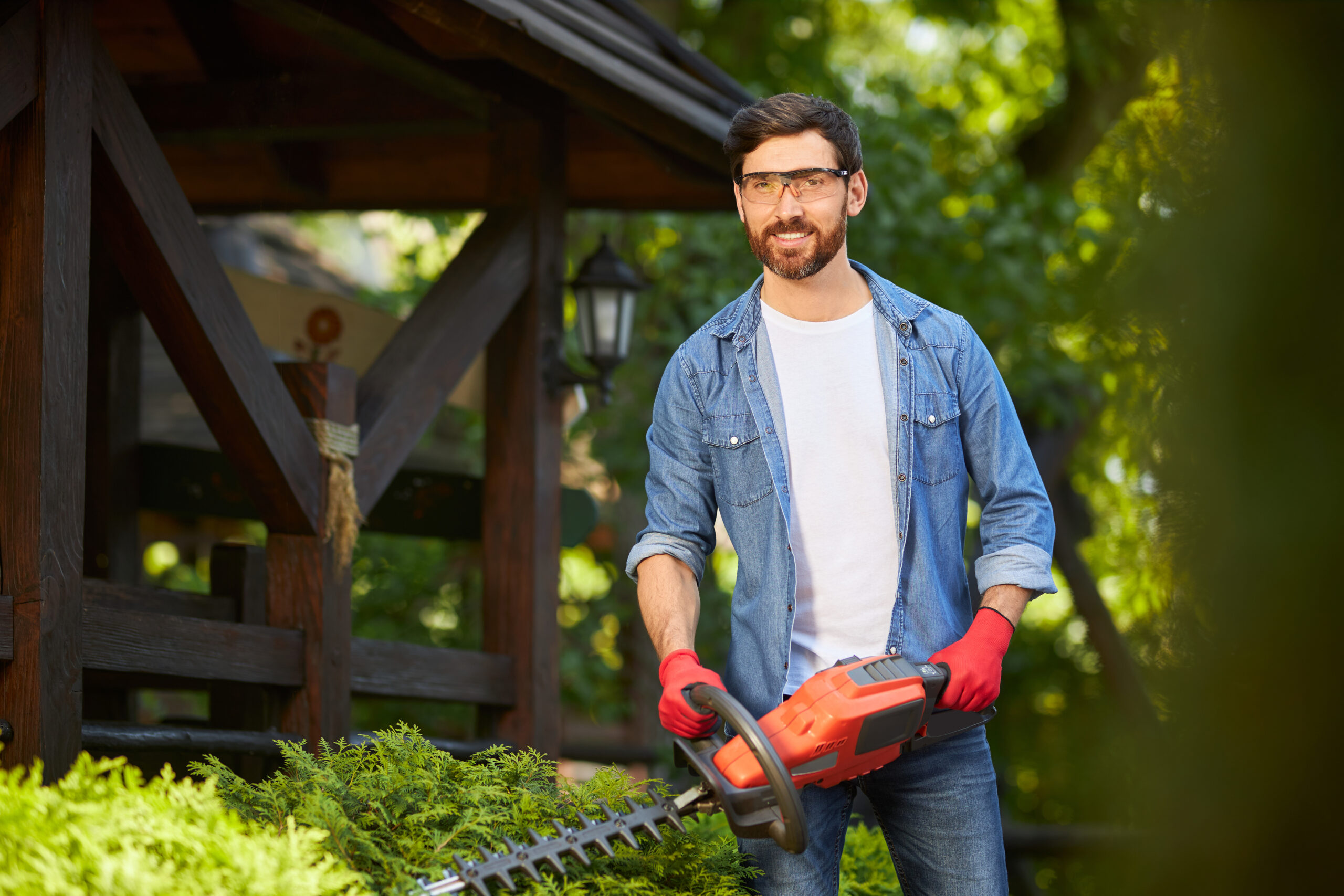 Attractive gardener posing, while trimming conifer bush by electric hedge clippers in sunny day. Portrait of male worker smiling at camera, while trimming thuja in garden. Concept of seasonal work.